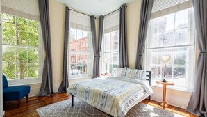 A bedroom with a bed, a blue chair, and large windows with curtains. - Bright & Sunny CBD FQ Convention Center Area Condo (New Orleans)