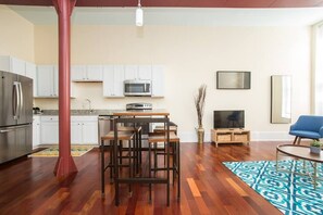 Cocina moderna con gabinetes blancos, microondas y refrigerador. Hay una zona de comedor con mesa y sillas, un televisor y una silla azul.
