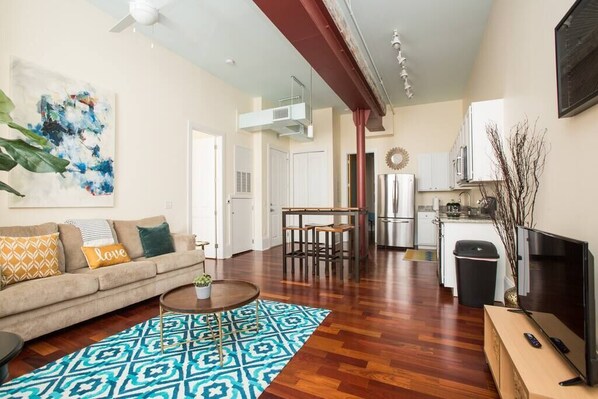 Una sala de estar con sofá, mesa de centro y un tapete estampado. Hay una zona de cocina con armarios y un refrigerador.