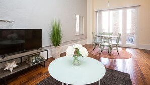 Ett vardagsrum med en platt-TV, ett runt, vitt bord med en vas med blommor, en krukväxt och en matplats med stolar och ett bord.