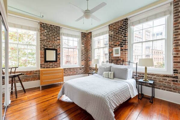 Quarto com parede de tijolos, cama com almofadas, cômoda, cadeira e janela com vista para prédios.