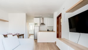 Ein modernes, helles Wohnzimmer mit einem weißen Sofa, einem Essbereich mit rosafarbenen Tellern und einer Küche mit weißen Schränken und einem eingebauten Fernseher.