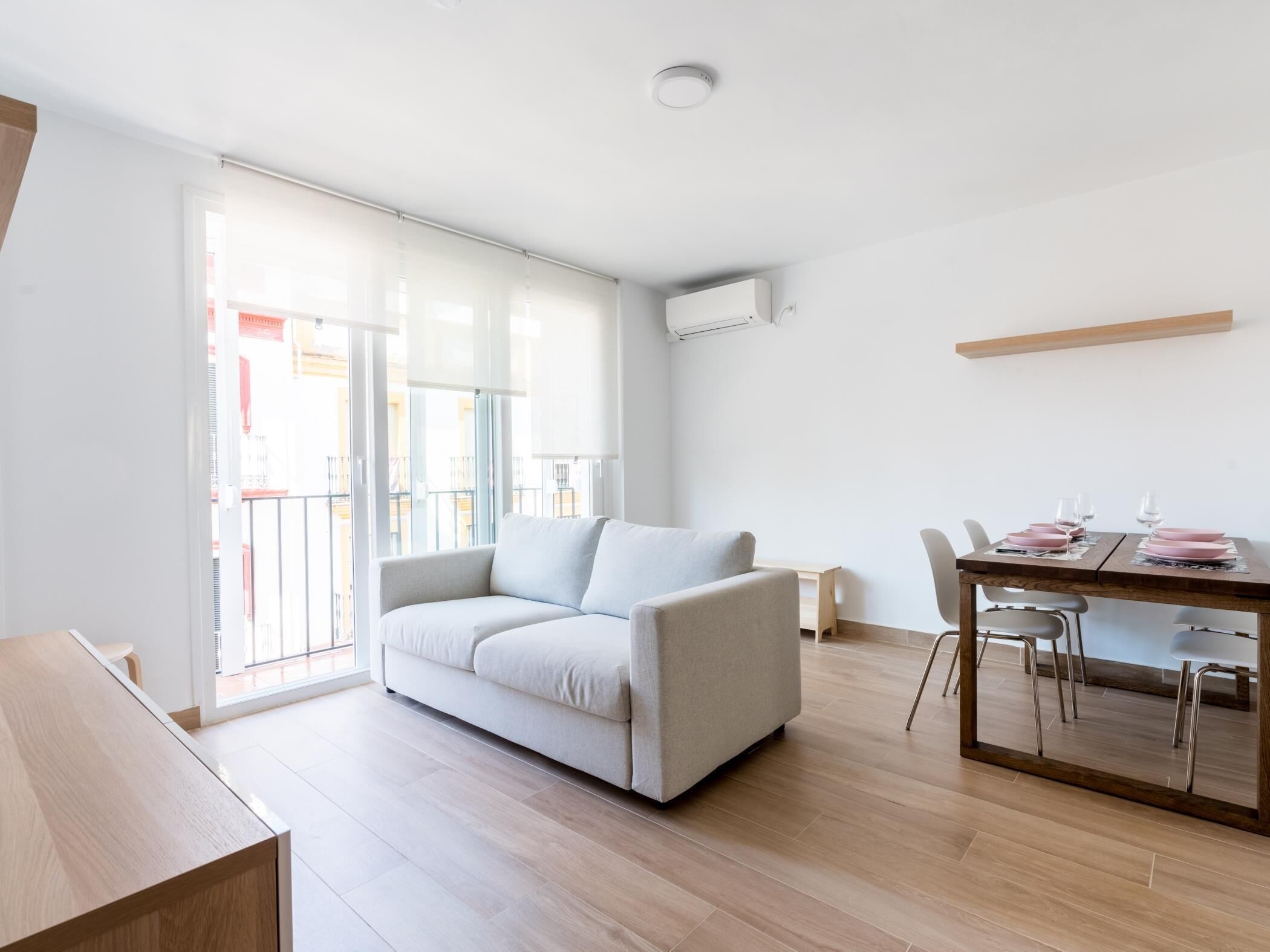 A modern living room with a sofa, a dining table set for two, and a sliding glass door leading to an outdoor balcony.