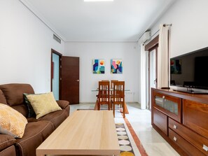 Una sala de estar moderna con un sofá de cuero marrón, una mesa de centro de madera y un comedor con sillas de madera y una mesa de vidrio. Hay pinturas abstractas en la pared, un televisor sobre un mueble de madera y una puerta con interruptor de luz.