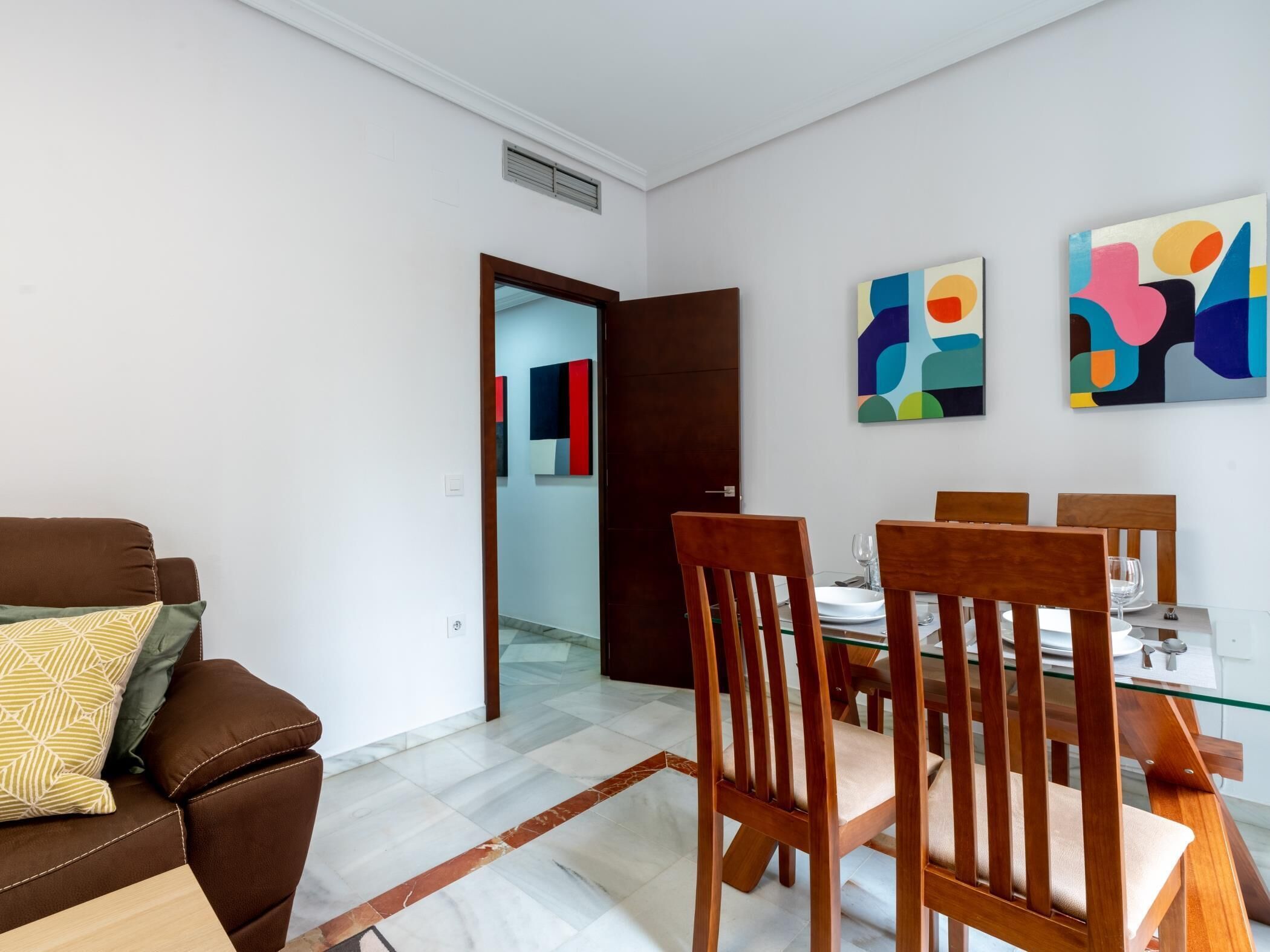 Un comedor moderno con una mesa de vidrio, sillas de madera y un sofá marrón con cojines estampados.