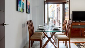 Een eethoek met een glazen tafel en houten stoelen, een televisie aan de rechterkant en kleurrijke abstracte schilderijen aan de muur.