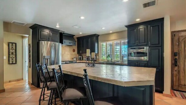 A kitchen with dark cabinetry, a large island with bar stools, stainless steel appliances, and a view of the outdoors.