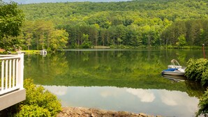 A serene lake with a boat docked on the right, surrounded by lush greenery and a forested hill in the background. - Lakefront, Kayaks, Firepit, great baseball stay! (Maryland)