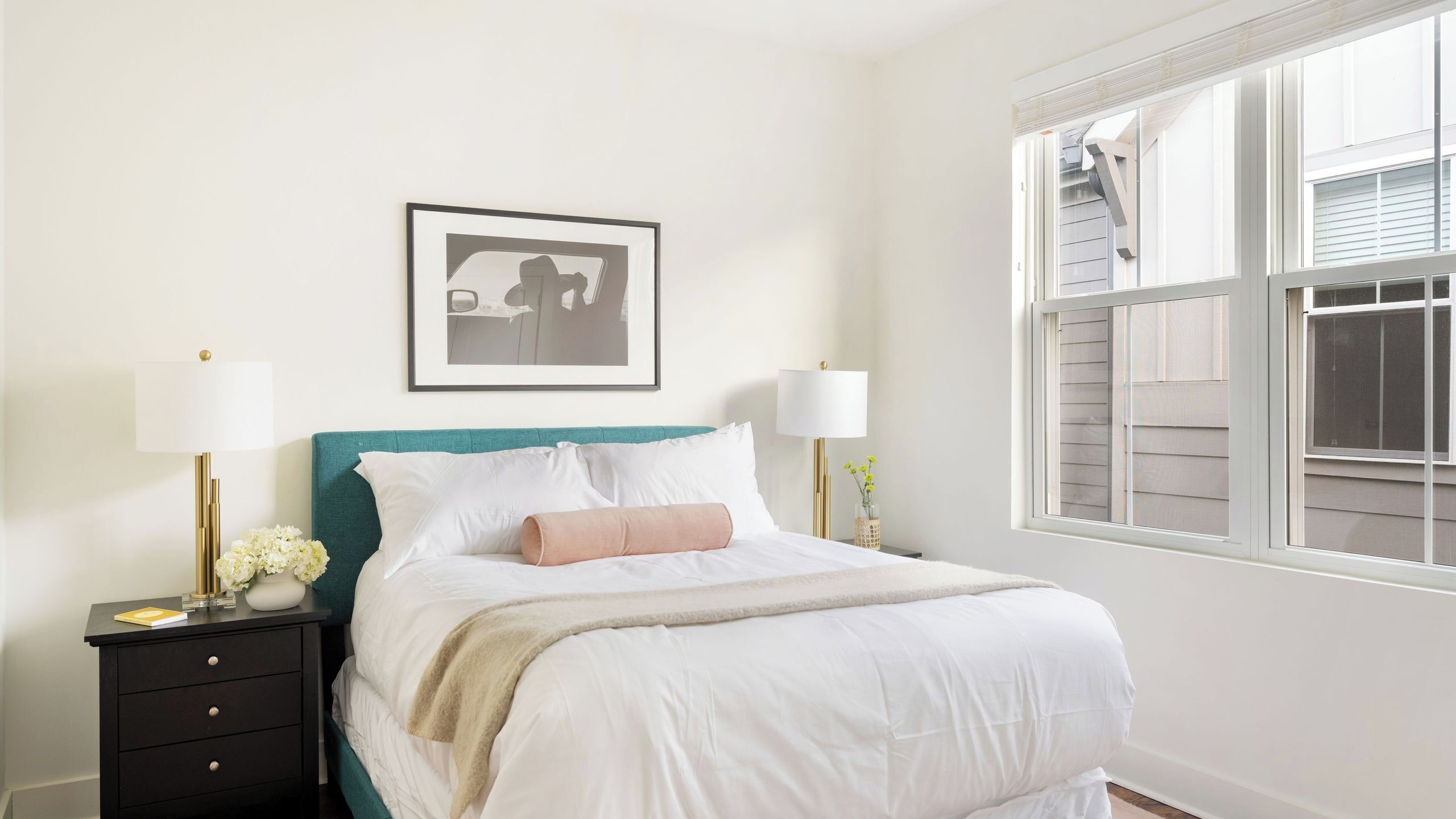 A neatly arranged bedroom with a bed, bedside lamps, a framed picture, and a window.