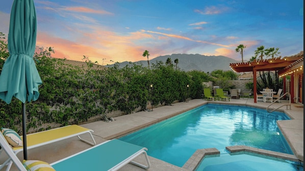 A swimming pool with lounge chairs, a pergola with lights, and a mountainous backdrop.