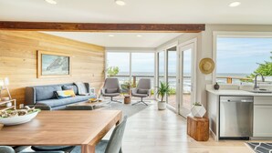 A modern living room with a wooden wall, a sofa, two chairs, a dining table, and a kitchen area with a view of the sea. - Sunset Cove by Avantstay Beachfront w/ Balcony! (Arch Cape)