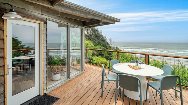 A wooden deck with a round table and chairs, overlooking a coastal view. - Sunset Cove by Avantstay Beachfront w/ Balcony! (Arch Cape)