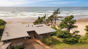 A modern house with a flat roof and a wooden deck, overlooking a beach and ocean. - Sunset Cove by Avantstay Beachfront w/ Balcony! (Arch Cape)