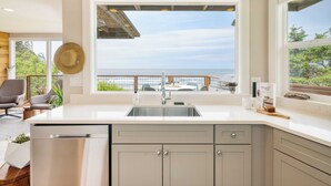 A modern kitchen with a large window overlooking the ocean, a built-in dishwasher, and a wooden dining area. - Sunset Cove by Avantstay Beachfront w/ Balcony! (Arch Cape)