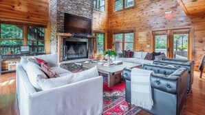 A spacious living area with a stone fireplace, comfortable seating, and large windows offering views of the outdoors. - Hickory Ridge by Avantstay Sleeps 20! Incredible Private Elevated Cabin (Gatlinburg)
