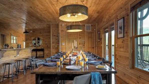 A wooden cabin with a long dining table set for a meal, bar stools, and large windows. - Hickory Ridge by Avantstay Sleeps 20! Incredible Private Elevated Cabin (Gatlinburg)