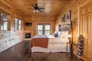A wooden cabin bedroom with a bed, a fireplace, a ceiling fan, and a view through the windows.