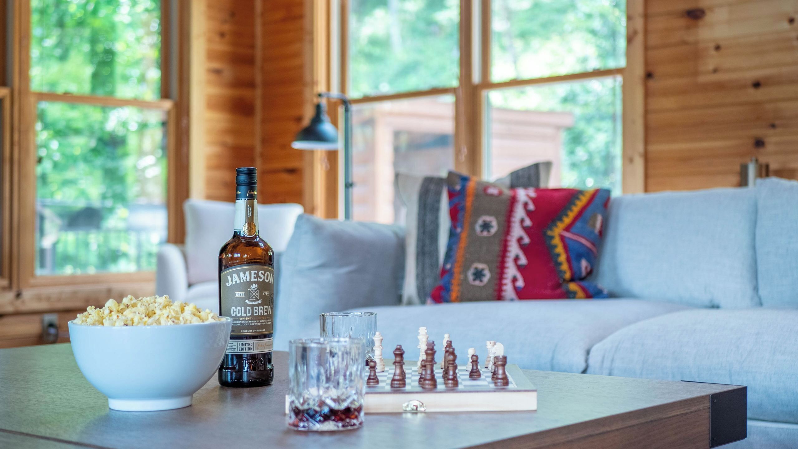 A cozy room with a wooden interior, a white sofa, a bowl of popcorn, a bottle of Jameson whiskey, a glass, and a chessboard.