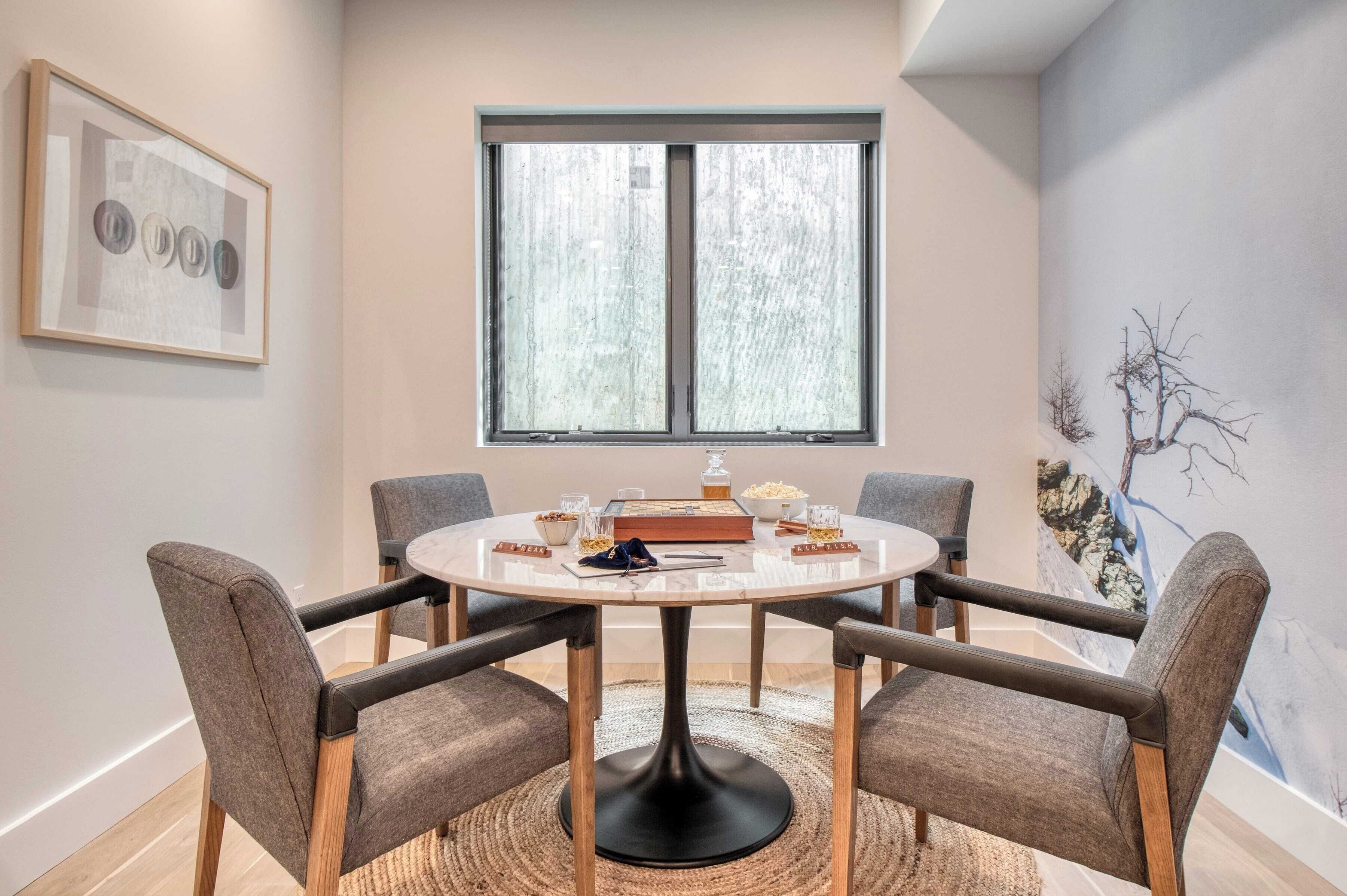 A dining area with a round table, four chairs, a large window, and a framed artwork on the wall.