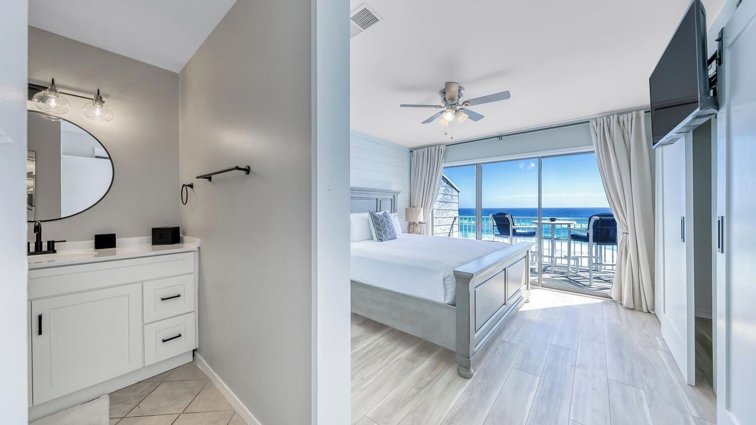 Un dormitorio moderno con una cama grande, ventilador de techo, balcón con vistas y baño con espejo e iluminación.