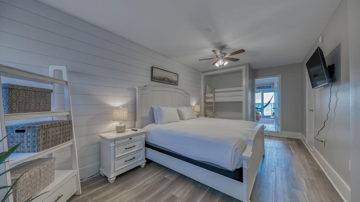 A bedroom with a bed, nightstands, a ceiling fan, a TV, and a view of the beach through a door.