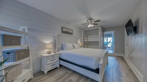 A bedroom with a bed, nightstands, a ceiling fan, a TV, and a view of the beach through a door.
