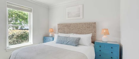 Un dormitorio con cama, ventilador de techo, ventana con vista a follaje y cabecera de mimbre, junto a dos mesitas de noche con lámparas.