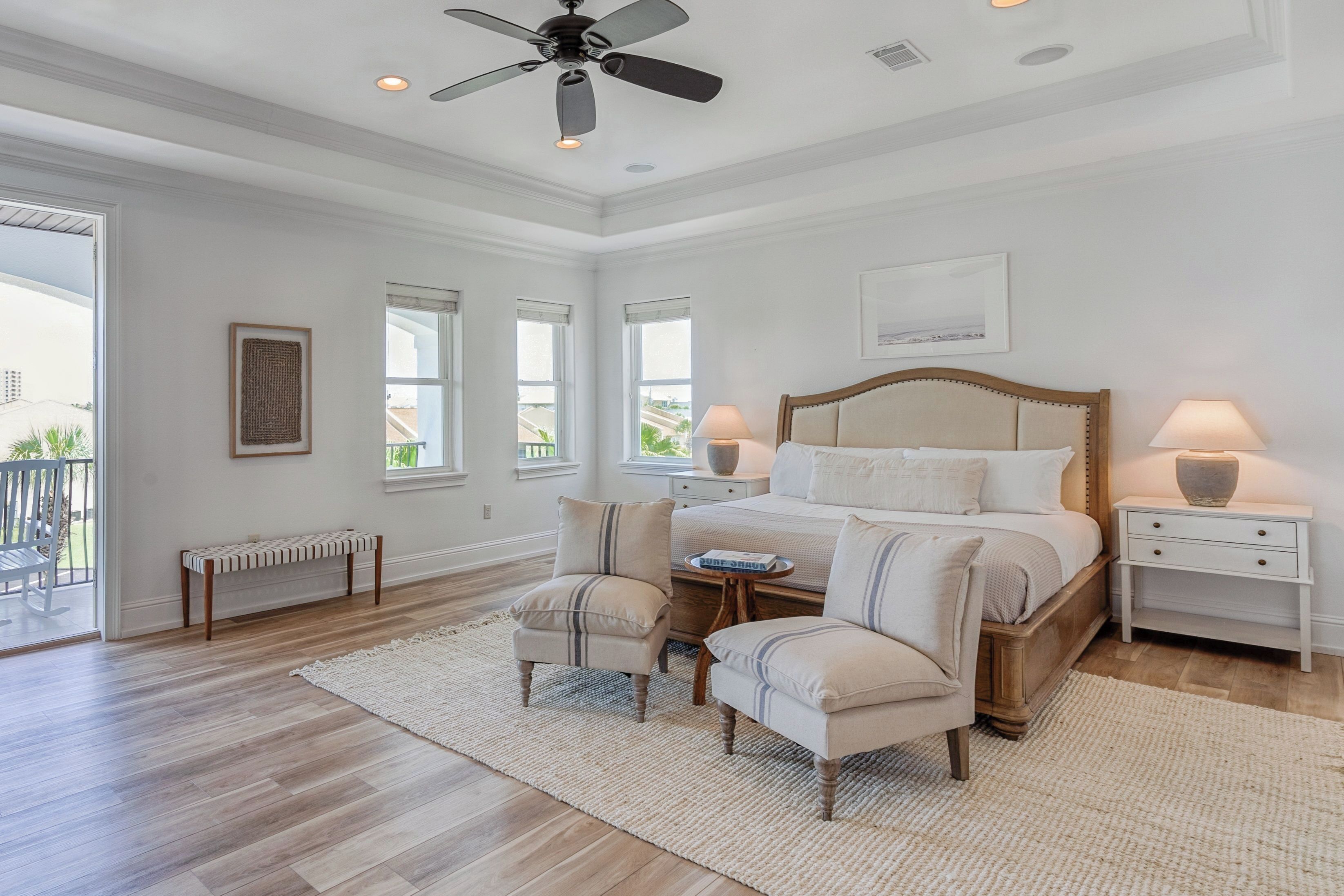 A bedroom with a large bed, two bedside tables, a ceiling fan, and a view of the outside.
