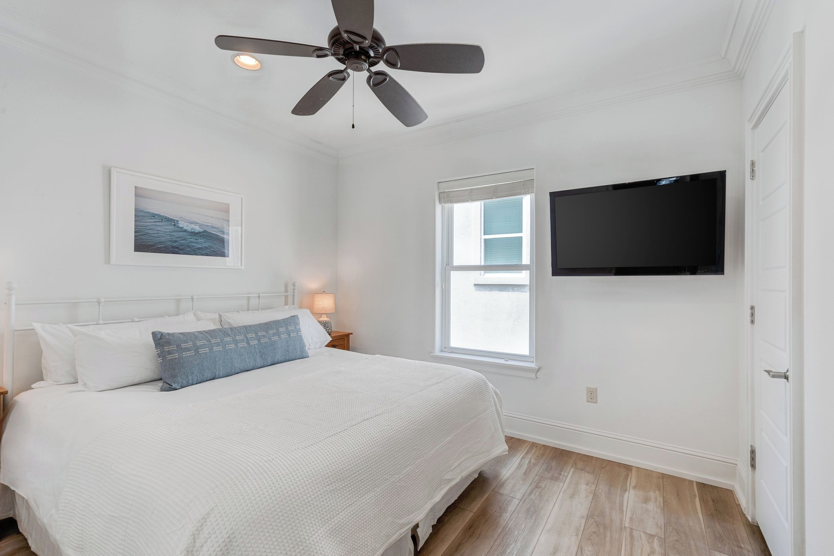 A bedroom with a bed, a ceiling fan, a TV, a window, and a nightstand with a lamp.