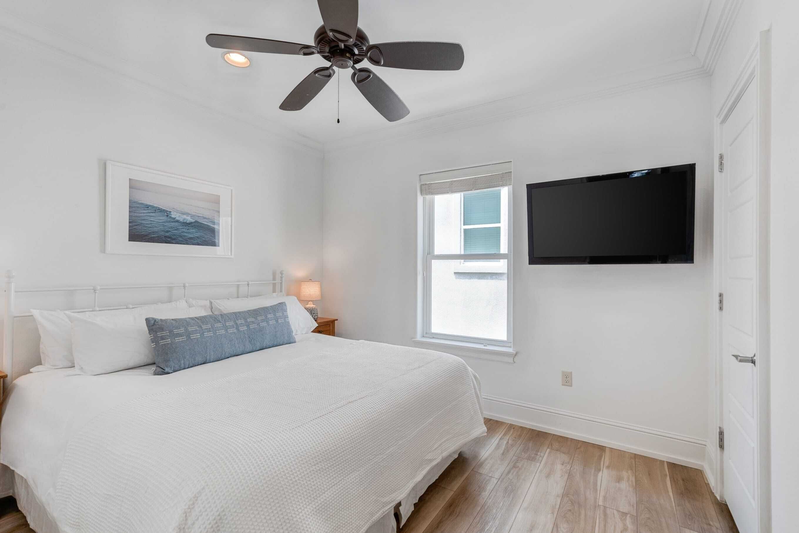 A bedroom with a bed, a ceiling fan, a TV mounted on the wall, a window, and a nightstand with a lamp.