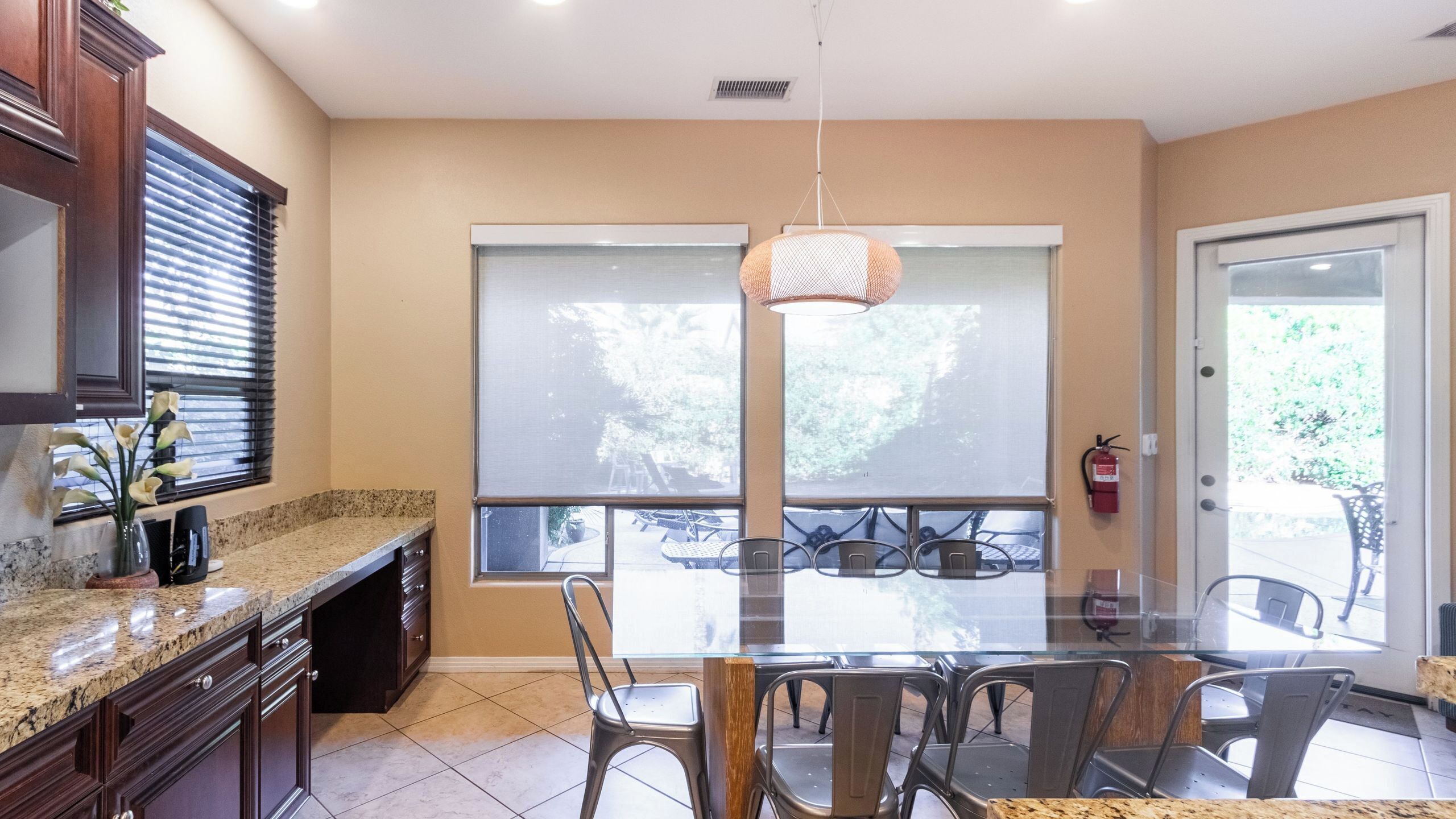 A kitchen with a granite countertop, dark wooden cabinets, a glass dining table with metal chairs, and a large window with blinds.