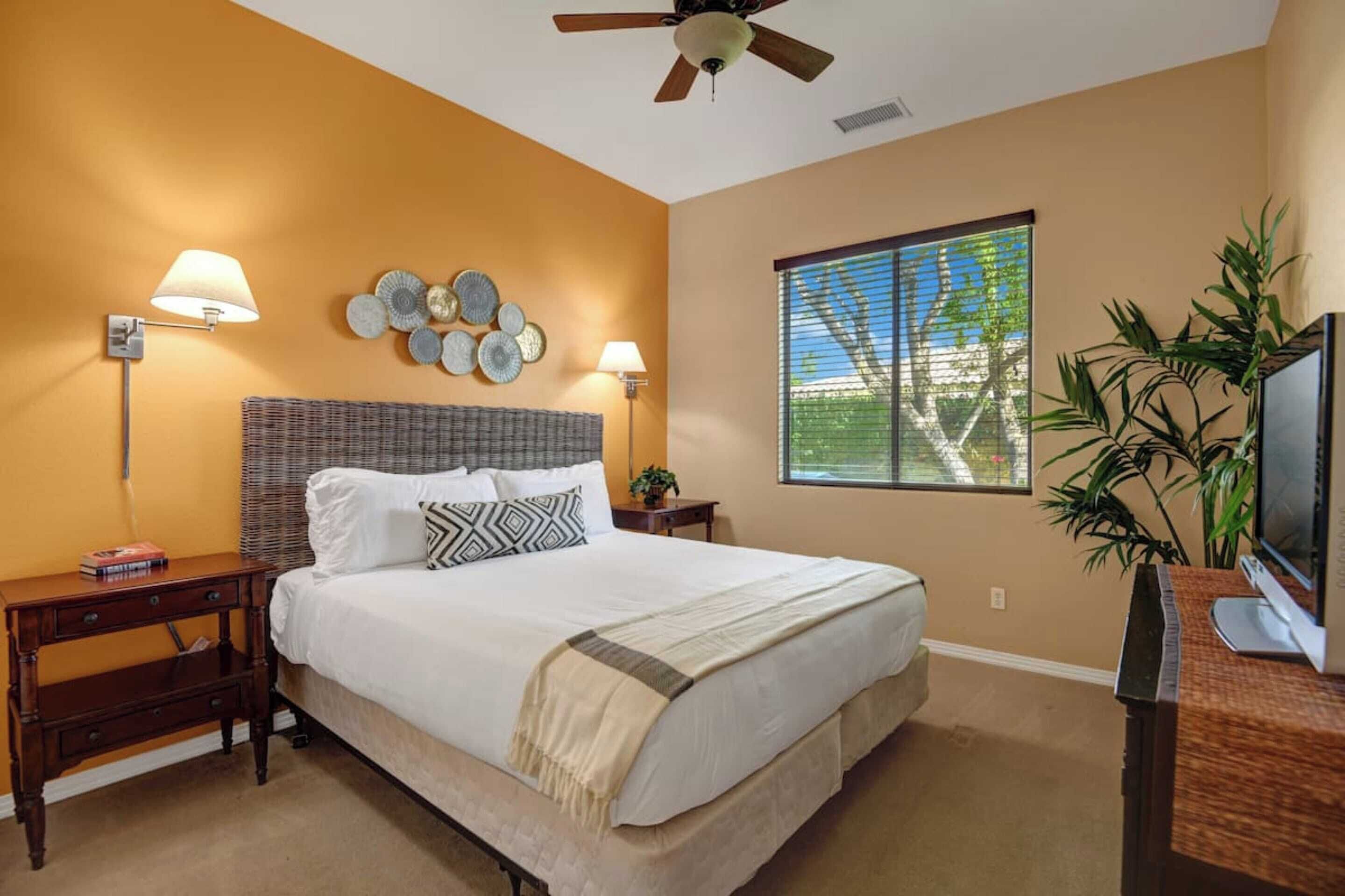 A bedroom with a large bed, two bedside tables, a ceiling fan, a window with blinds, and a television on a wooden stand.