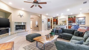 A spacious living room with a flat-screen TV, a fireplace, a dining area, and a kitchen with wooden cabinets. - Songbird by Avantstay Pool, Spa & Firepit + Tons of Outdoor Seating (Indio)