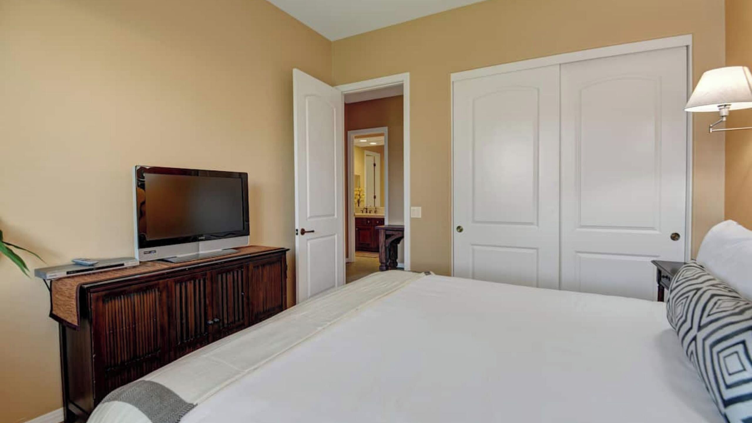 A hotel room with a bed, a television on a wooden dresser, a lamp, and a door leading to a bathroom.
