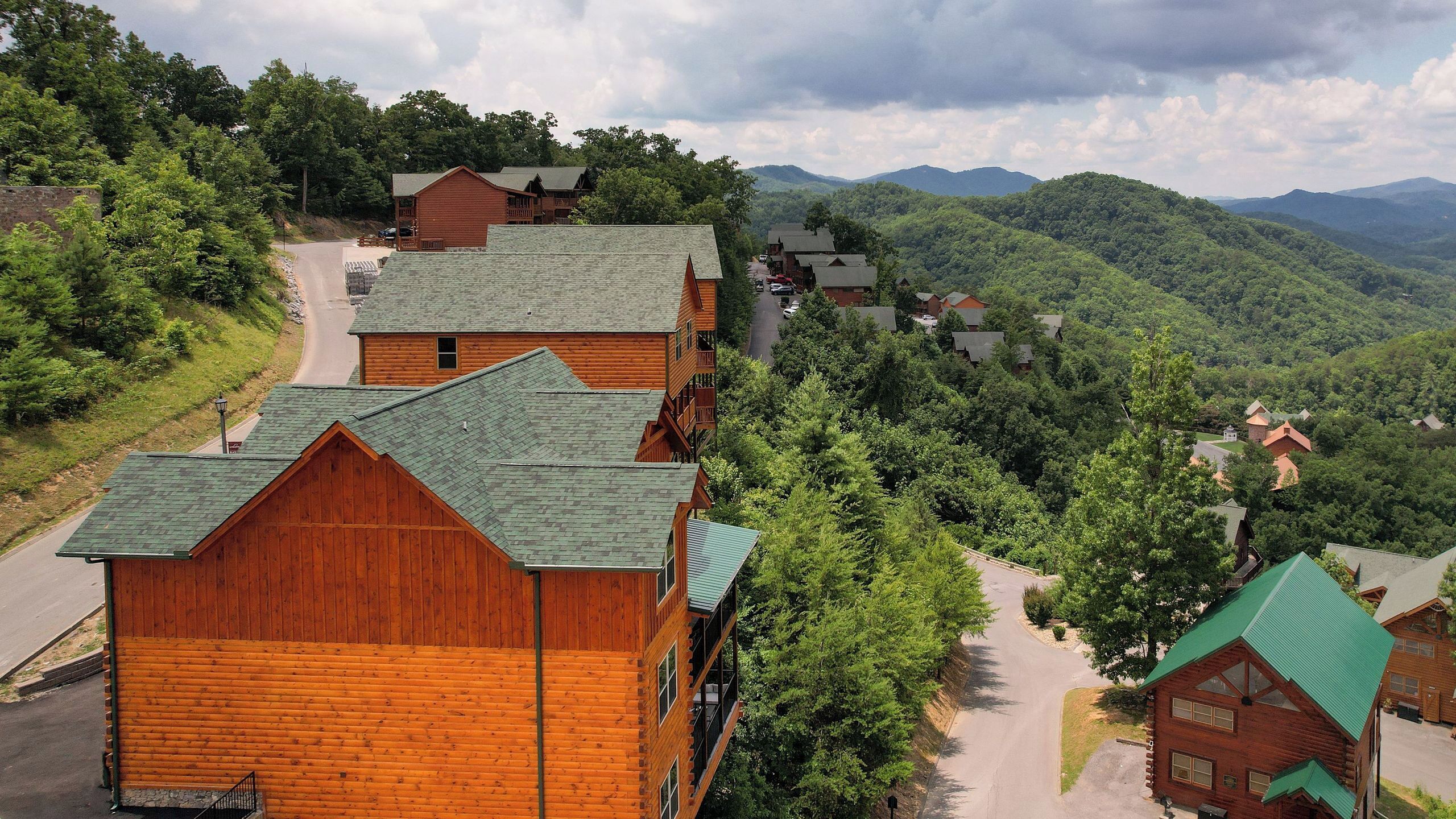 A cluster of wooden cabins with green roofs, surrounded by lush greenery and mountains in the background.