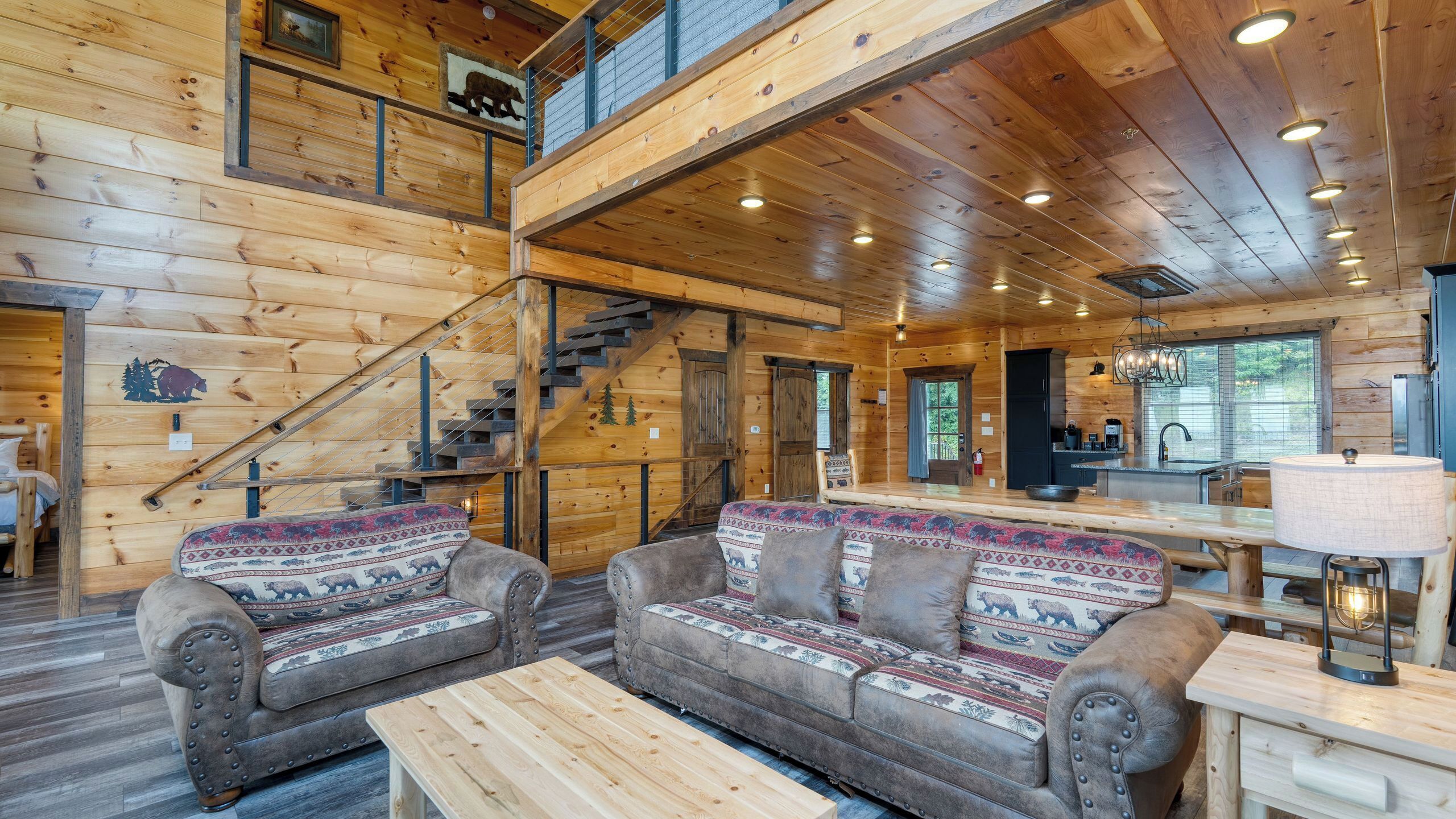 A log cabin interior with a staircase, two patterned sofas, a wooden table, and a kitchen area.