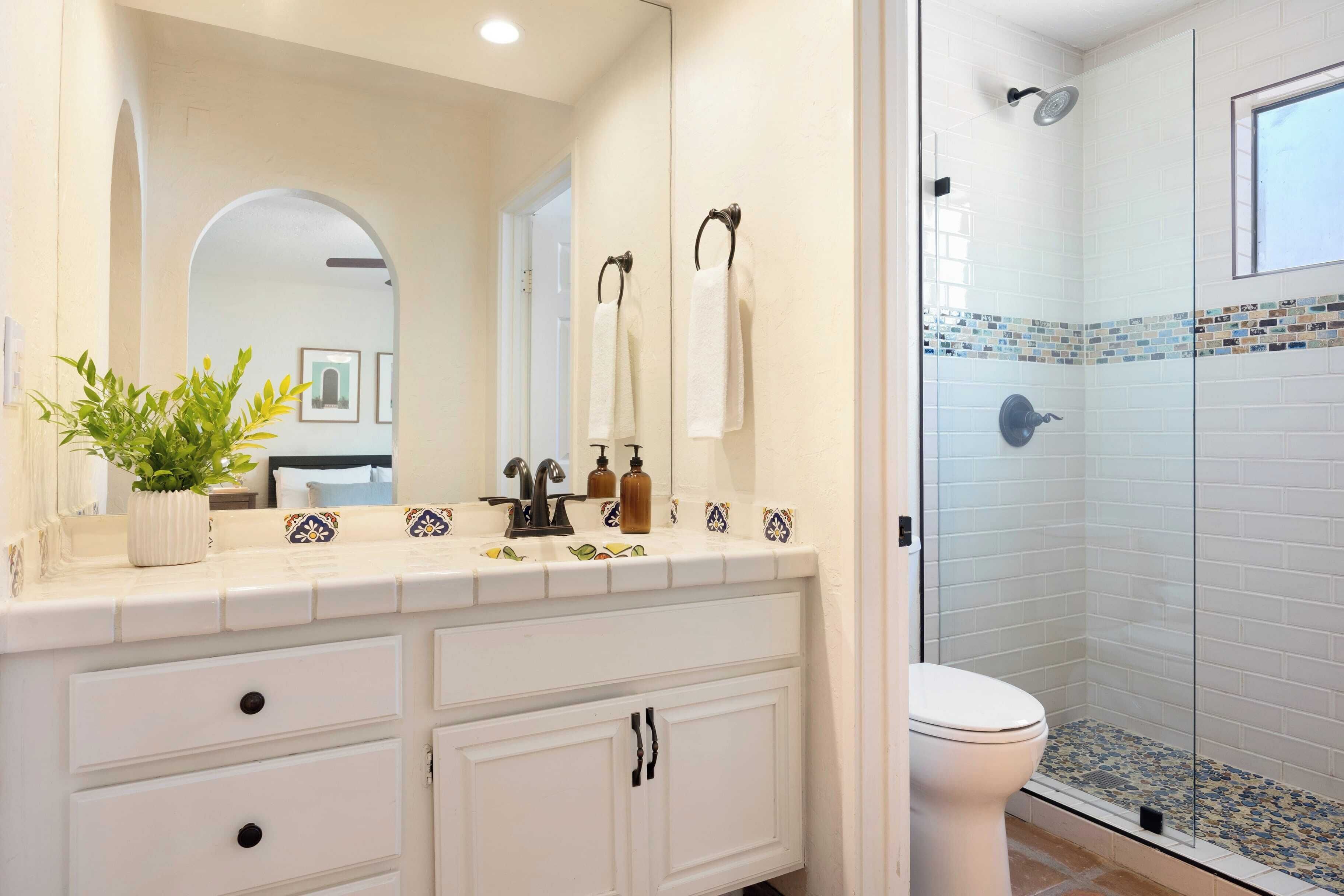 Une salle de bain avec un meuble-lavabo blanc, un miroir, une plante en pot et un espace douche orné de carreaux de mosaïque.