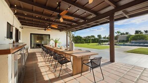 An outdoor patio with a wooden table and chairs, a bar area, ceiling fans, and a view of a landscaped garden. - Sleeps 20! Architectural Mansion w/ Pool, Indoor Tennis Court, Game Room Starlight Estate by Avantstay (Indio)