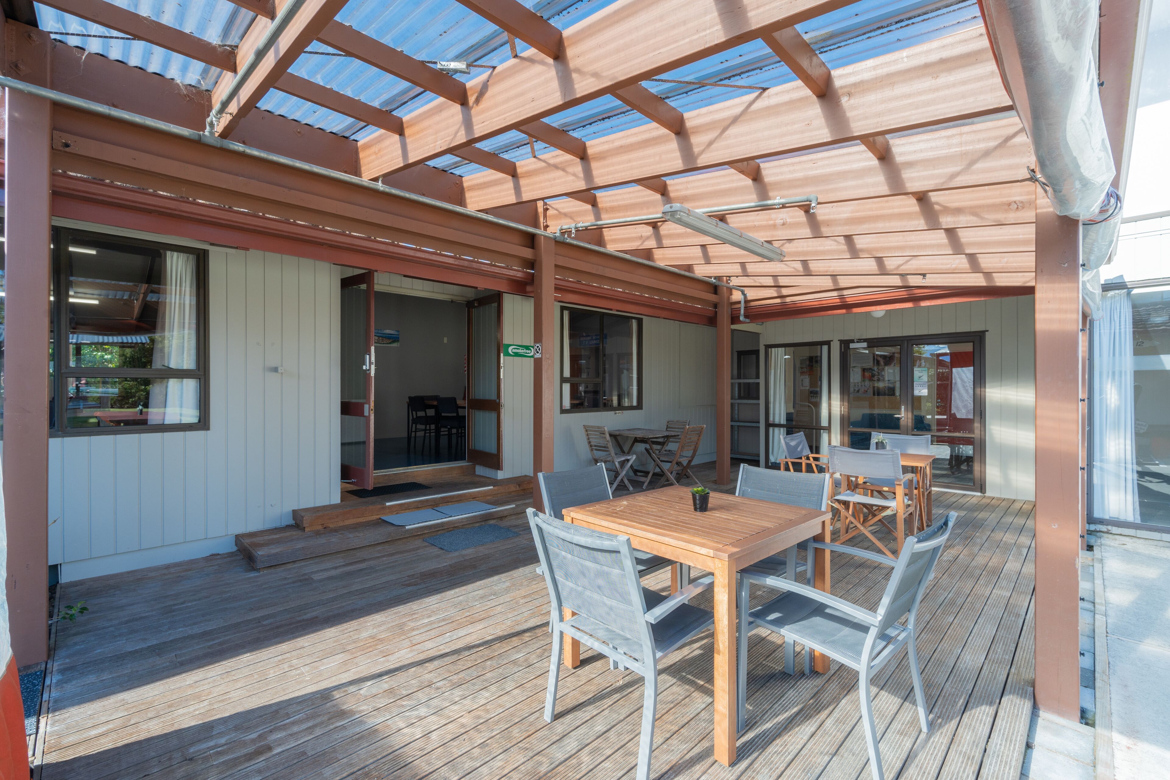 A covered outdoor dining area with a wooden table and chairs, a glass door entrance, and a view of the adjacent building.