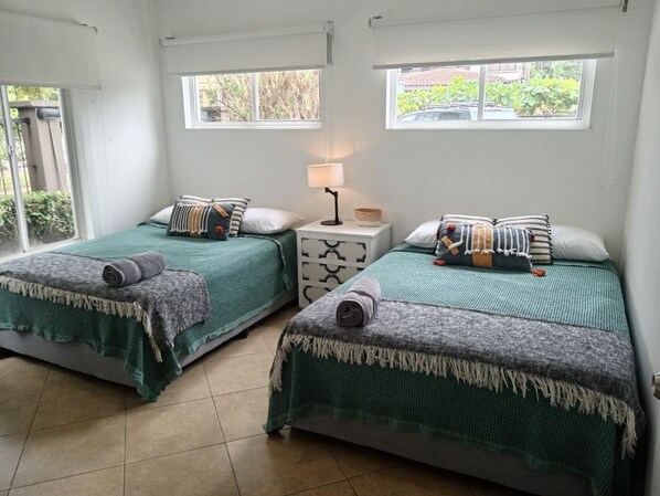 A room with two beds, each with a green bedspread and a rolled towel. There is a nightstand with a lamp between the beds.
