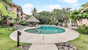 Un espace piscine au design incurvé, doté de chaises longues, d’une structure à toit de chaume et d’une végétation tropicale.