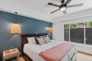 Un dormitorio con una cama, un ventilador de techo, dos lámparas de pared, un ventanal y una mesita con una planta.