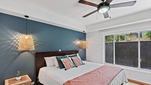 Une chambre à coucher avec un lit, deux lampes fixées au mur, un ventilateur de plafond et une fenêtre donnant sur un paysage verdoyant.