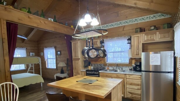 A cozy cabin-style kitchen with wooden floors, a central island with stools, a hanging light fixture, and a refrigerator. - A Peace of Heaven LOG Cabins & RV- Cabin #1 (Vanderpool)