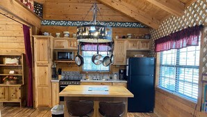 A rustic kitchen with wooden cabinets, a black refrigerator, a wooden island with bar stools, and hanging pots. - A Peace of Heaven LOG Cabins & RV- Cabin #3 (Vanderpool)