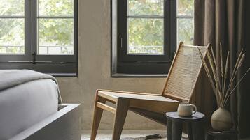 A room with a large arched window, a wooden chair, a small table with a mug, and a vase with reeds.