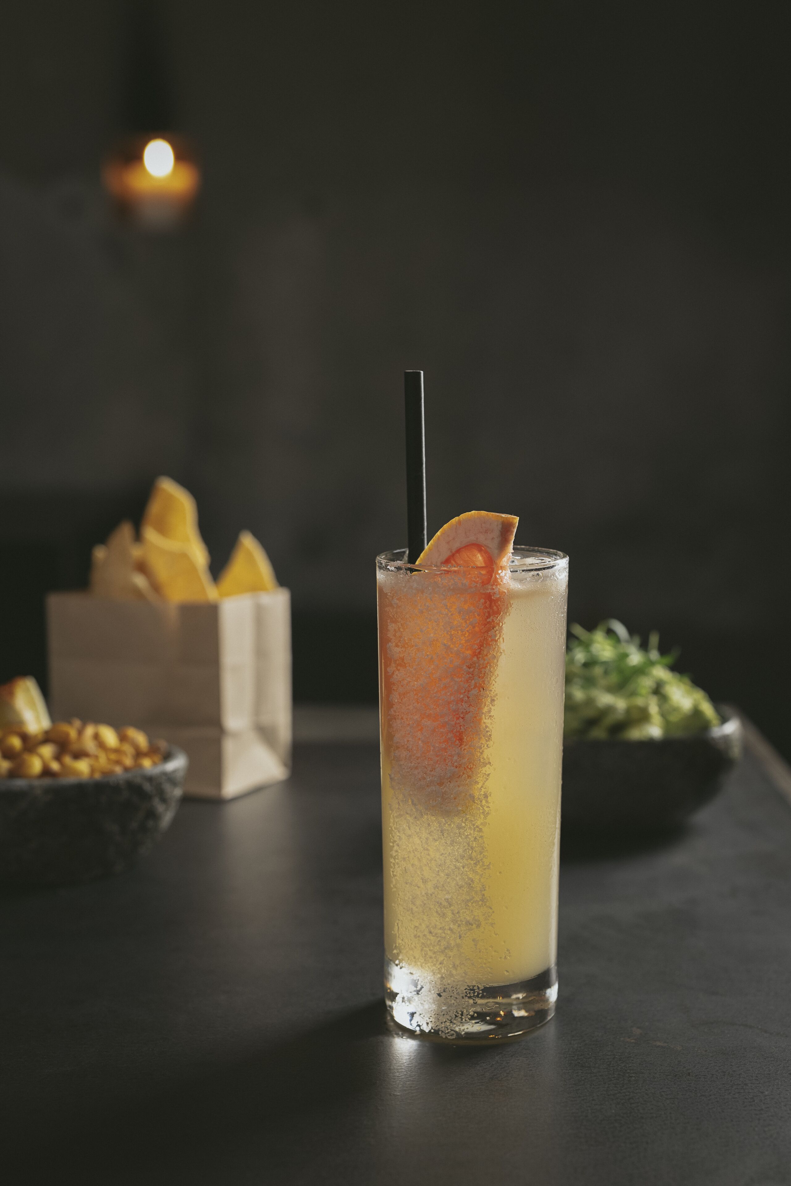 Un grand verre avec une paille, contenant une boisson jaune et décoré d’une tranche d’orange, posé sur une surface sombre.