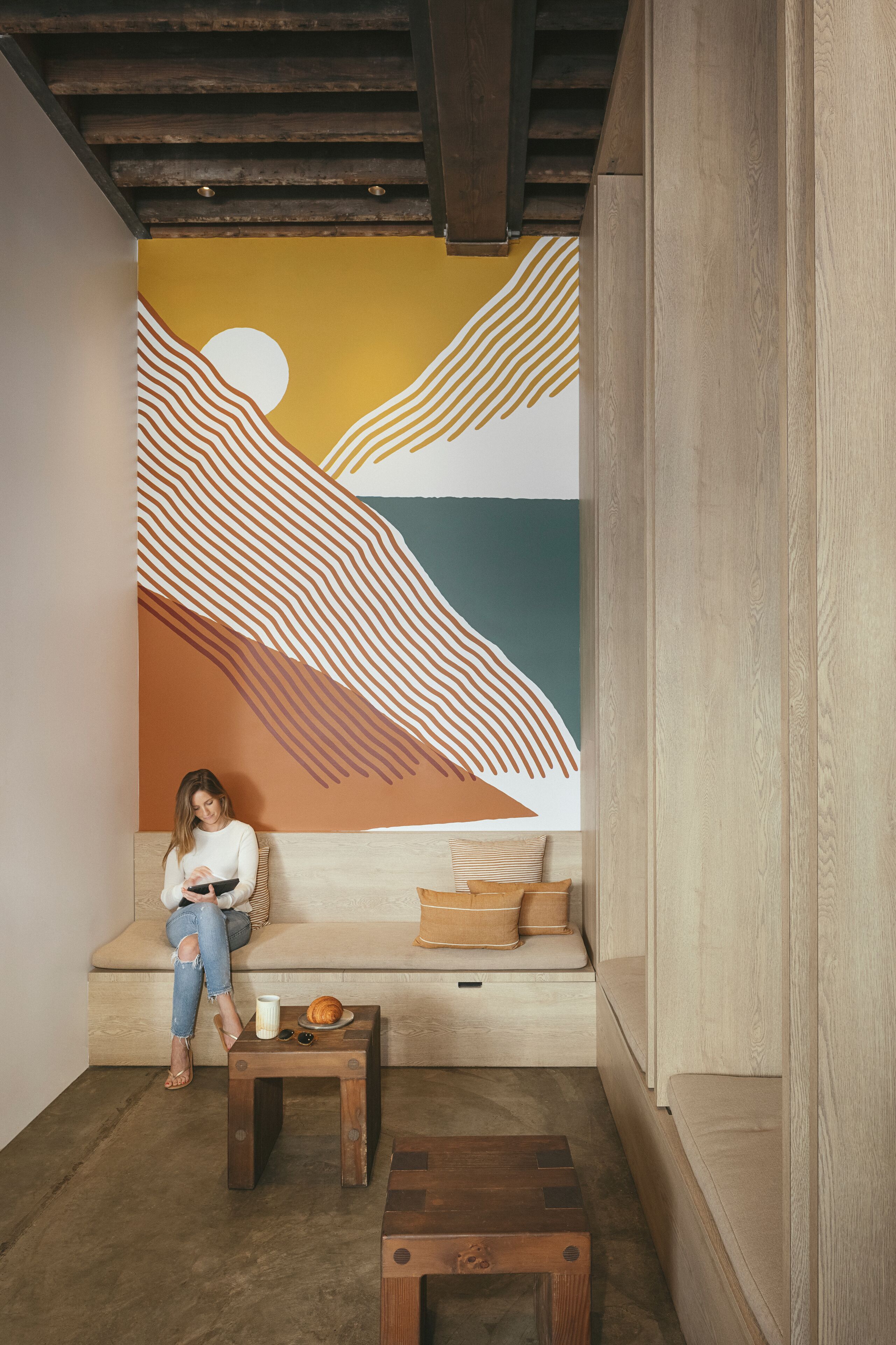 A cozy seating area with a mural, a wooden bench, and a small table with a croissant and sunglasses.