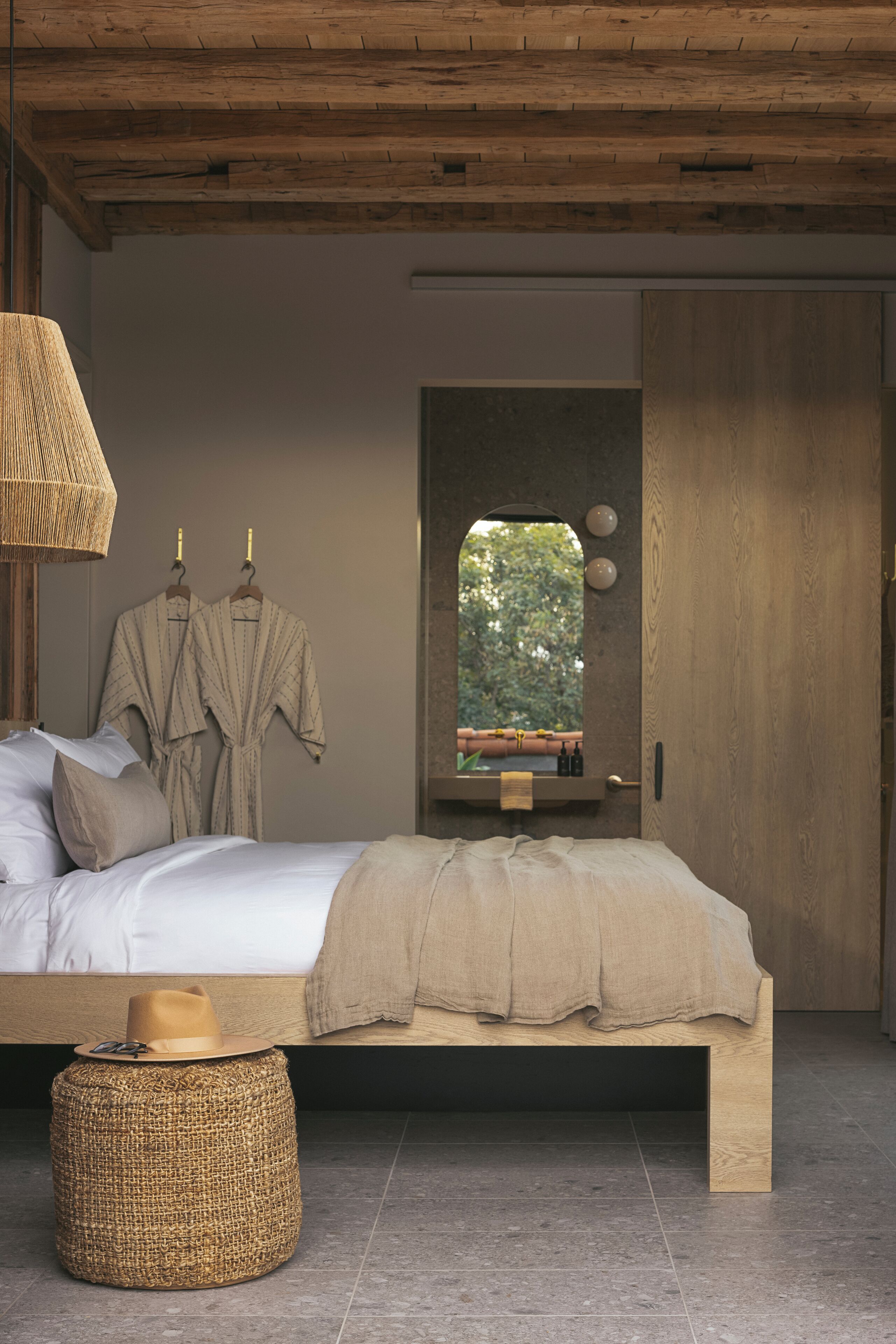 A modern bedroom with a wooden ceiling, a large bed with a headboard, a bedside table with a lamp, and a mirror above the door.