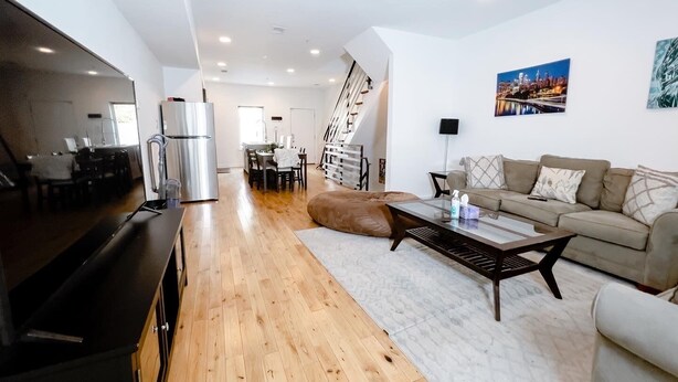 A modern living room with a sofa, coffee table, and a TV. There is a kitchen area with a refrigerator and a dining table. A staircase leads to another level. - Fantastic 4BR Townhouse W/ HOTTUB (Philadelphia)